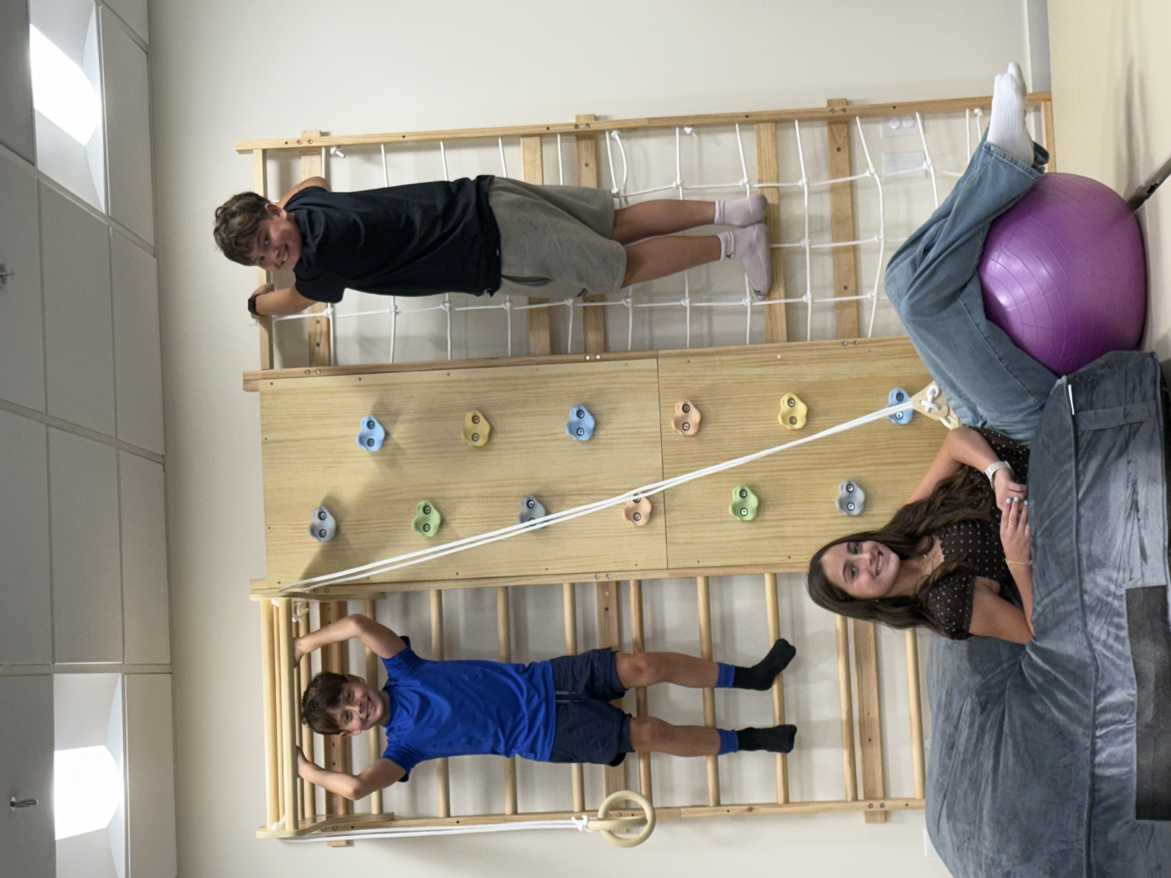 Three kids enjoying gross motor play on the climbing wall and monkey bars in the Mighty Miracles Therapy gym.