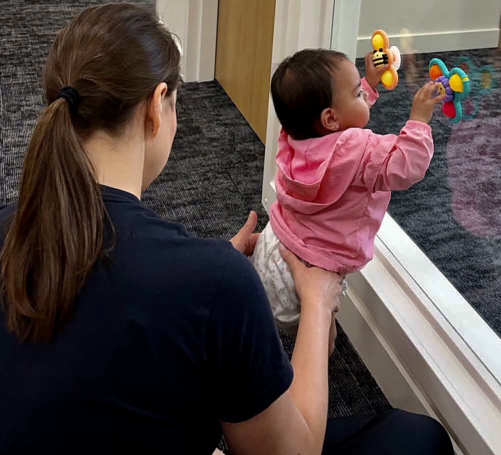 A therapist holds a toddler in a pink jacket up to a window, helping her reach for colorful hanging toys during a play session.