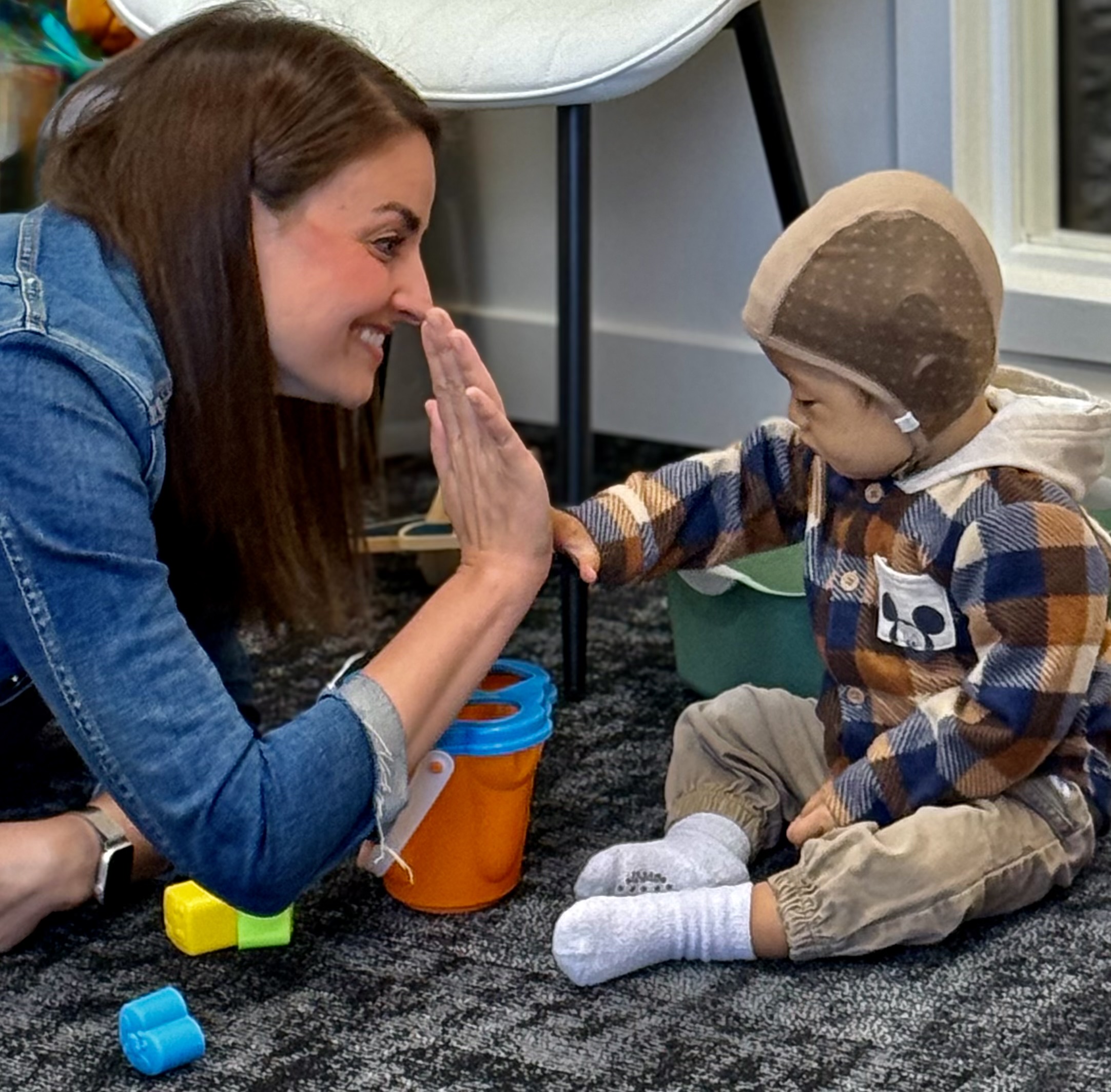 A toddler plays with a bright yellow cup while sitting on a soft rug, with two adults encouraging her exploration in a warm, inviting atmosphere.